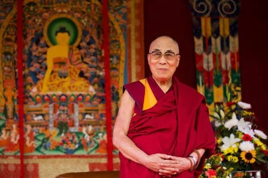 A Tibetan Buddhist teacher in maroon robes stands before a thangka painting of the Buddha